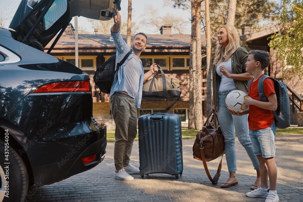 Happy young family with little packing stuff into the car and smiling while standing near house