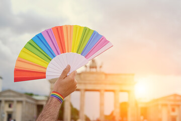 Rainbow fan in hand, symbol of LGBTQIA gay lesbian pride. Rainbow also symbolizes diversity in covid dissident movement, querdenken in German. Sunset over Brandenburg Gate in Berlin, Germany.