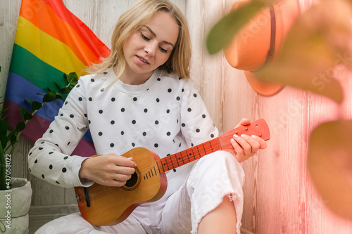 Young millennial hippie woman sitting on balcony play guitar. Music lesson and singer sitting. LGBTQ rainbow flag on background. Online music tuition. Playing instrument