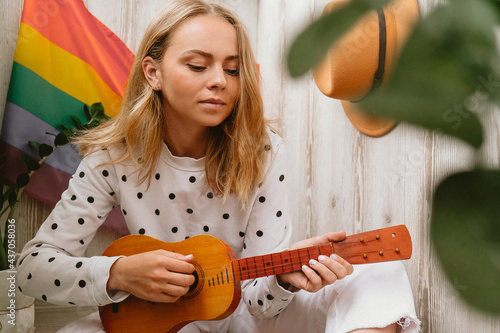 Young millennial hippie woman sitting on balcony play guitar. Music lesson and singer sitting. LGBTQ rainbow flag on background. Online music tuition. Playing instrument