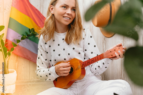 Young millennial hippie woman sitting on balcony play guitar. Music lesson and singer sitting. LGBTQ rainbow flag on background. Online music tuition. Playing instrument