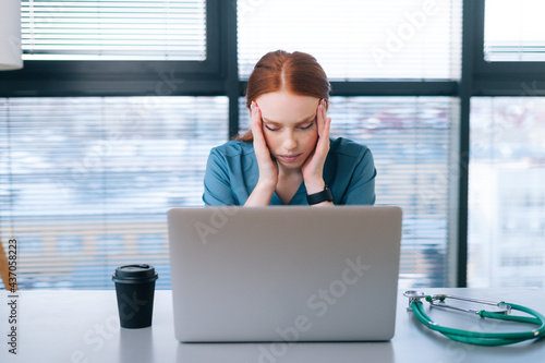 Side view of tired exhausted young female doctor in medical uniform having headache and rubbing head while working on laptop sitting at desk in office of medic clinic.