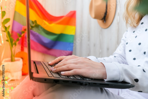 Young millennial hippie woman sitting on balcony using laptop. LGBTQ rainbow flag on background. Online education tuition. Work from home. Modern office.