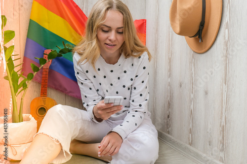 Young millennial hippie woman sitting on balcony using mobile phone. LGBTQ rainbow flag on background. Phone texting, video call, taking selfie. Blogger. Work from home.