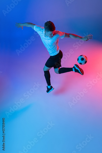 High angle view of young man, male soccer football player training isolated on gradient blue pink background in neon light.