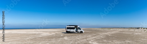 panorama of a gray camper van parked on an endless white sand beach in the middle of nowhere with ocean behind