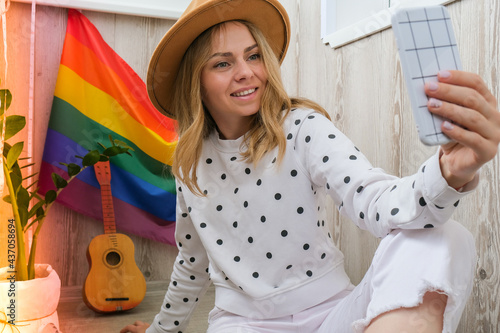 Young millennial hippie woman sitting on balcony using mobile phone. LGBTQ rainbow flag on background. Phone texting, video call, taking selfie. Blogger. Work from home.