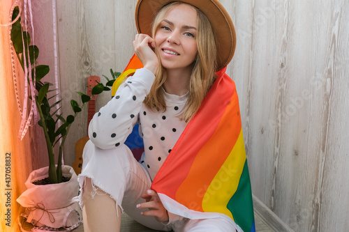 Young blonde millennial hippie woman with rainbow LGBTQ flag at home balcony. Peace and freedom the symbol of LGBT, spending and sharing loving time. Support LGBTQ community.