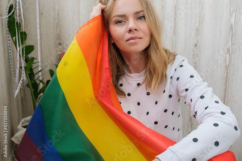 Young blonde millennial hippie woman with rainbow LGBTQ flag at home balcony. Peace and freedom the symbol of LGBT, spending and sharing loving time. Support LGBTQ community.