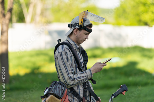 a young man in a protective plastic mask and with a trimmer on his shoulder holds a phone in his hands to answer a message or turn on music