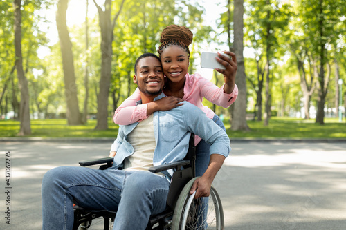 Happy black woman and her impaired boyfriend in wheelchair taking selfie together, hugging at city park