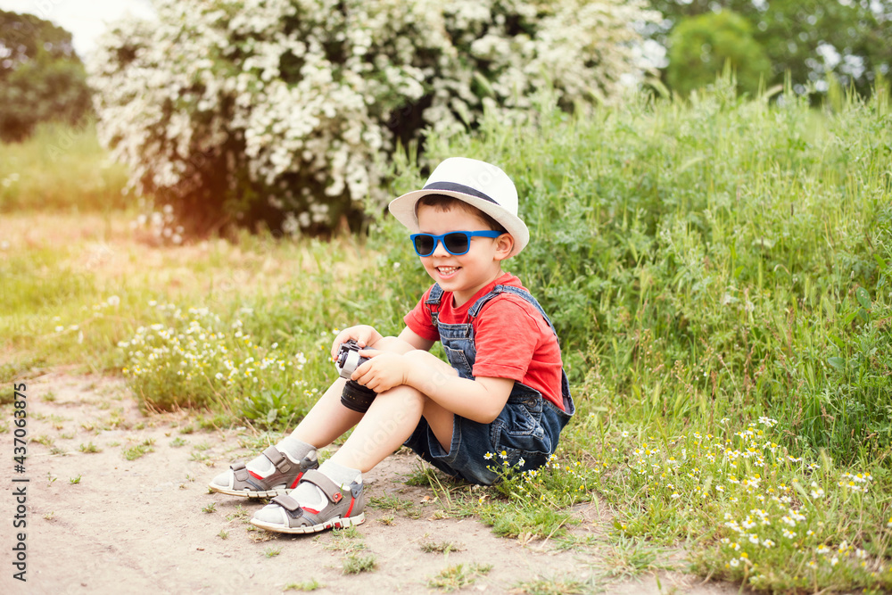 A cute little boy in denim overalls, blue sunglasses and a white hat sits on the grass in the park and holds an old retro camera in his hands. A cute little boy in denim overalls, blue sunglasses and a white hat sits on the grass in the park and holds an old retro camera in his hands.