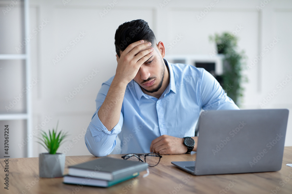 Tired millennial Arab businessman suffering from headache at desk in modern office Tired millennial Arab businessman suffering from headache at desk in modern office