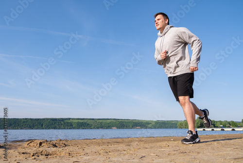 A young man in a gray sweatshirt runs on an empty beach at sunrise. Slimming and healthy lifestyle concept.