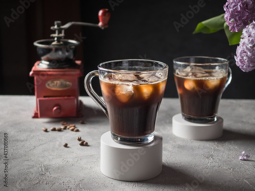 Black Iced Coffee in glass mugs with a round white plaster base. Iced coffee on a gray table. In the background are lilac flowers, a coffee grinder and coffee beans. Dark background.
