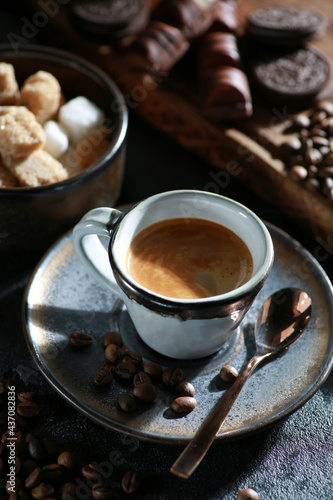 Morning coffee. Espresso in a small cup with a spoon on a saucer. Sugar, cookies, chocolate, coffee beans on a wooden board on a black table. Sunlight. Background image, copy space © Nadia