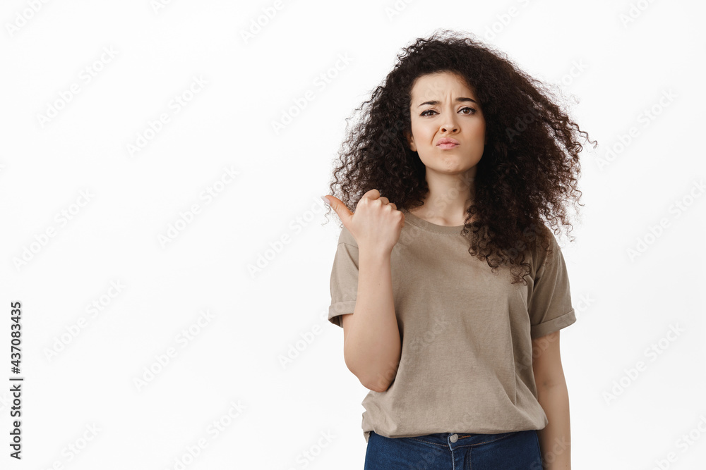 Disappointed young woman grimacing displeased, complaining on bad promo offer, pointing left at advertisement with upset skeptical face, standing over white background Disappointed young woman grimacing displeased, complaining on bad promo offer, pointing left at advertisement with upset skeptical face, standing over white background