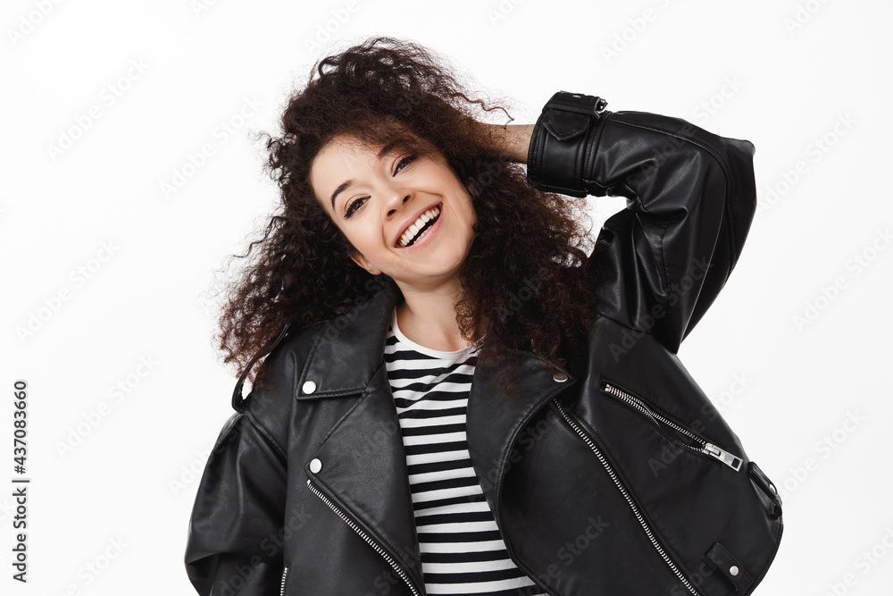 Close up of happy brunette girl with curly natural hair, smiling white teeth, touching stylish hairstyle and look carefree at camera, wearing leather jacket for summer evening walk, white background Close up of happy brunette girl with curly natural hair, smiling white teeth, touching stylish hairstyle and look carefree at camera, wearing leather jacket for summer evening walk, white background