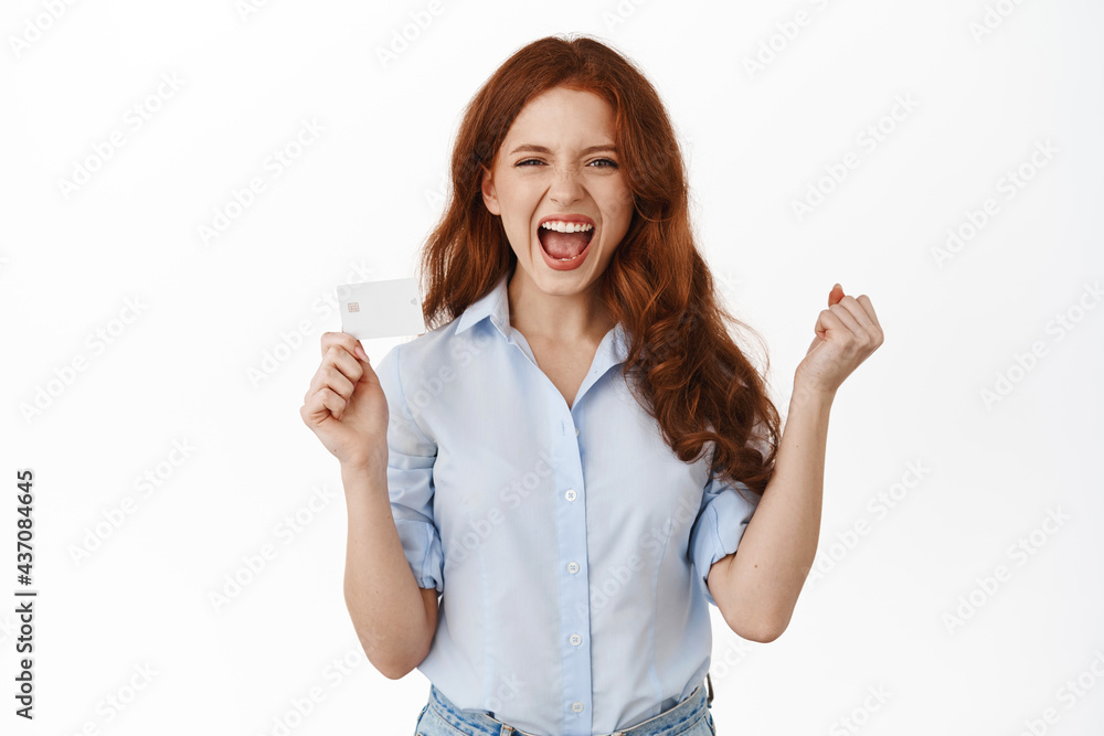 Cheerful redhead woman shouting with satisfaction, say yes and show credit card, ready for shopping maraphon with discounts, got new banking card, standing against white background Cheerful redhead woman shouting with satisfaction, say yes and show credit card, ready for shopping maraphon with discounts, got new banking card, standing against white background