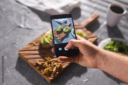 Hands take pictures on smartphone of two beautiful healthy sour cream and avocado sandwiches lying on board on the table. Social media and food concept