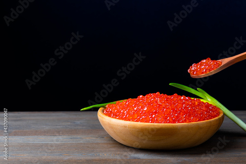 Red caviar in a wooden cup on a black background with a spoon and green onions. A large pile of bright caviar. Fresh delicious caviar. A place for advertising, logo, label, layout, mockup.
