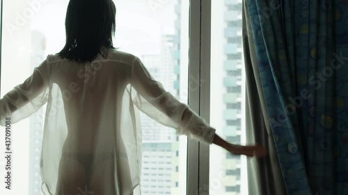 A woman wearing shear lingerie opens the bedroom curtains with optimism for opportunity in the new day