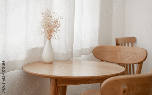 White vase with small flowers place on wooden table next to the window with white curtains and warm sunlight. Cozy and minimal room design