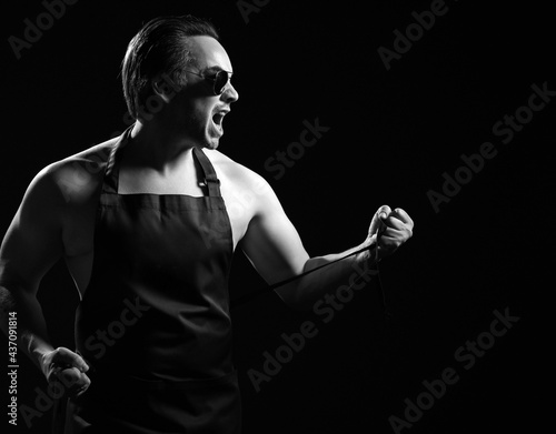 Black and white portrait of yelling loudly adult man butcher, farrier, barista in sunglasses and apron on naked body, looking aside at copy space body over black background