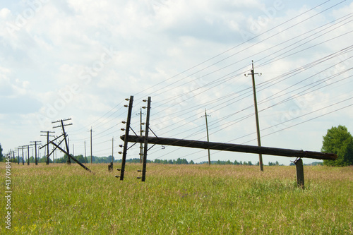 Two wooden poles of the power line broke and fell. The communication line goes into the distance. Supports stand in a blooming field. In the background, a cloudy sky, forest and concrete pillars 