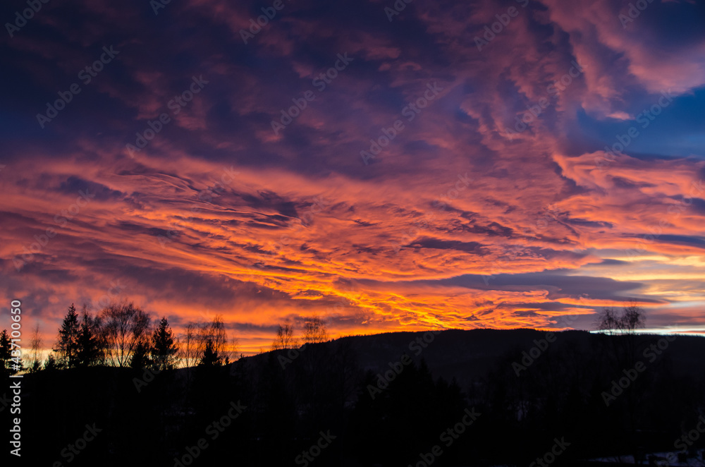 Sunset fire in the sky with multi color details and large space, tops of trees at the bottom and a mountain ridge in the distance