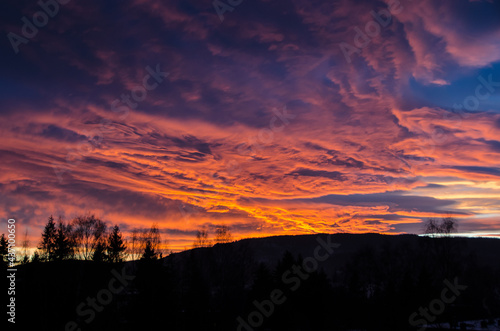 Sunset fire in the sky with multi color details and large space  tops of trees at the bottom and a mountain ridge in the distance