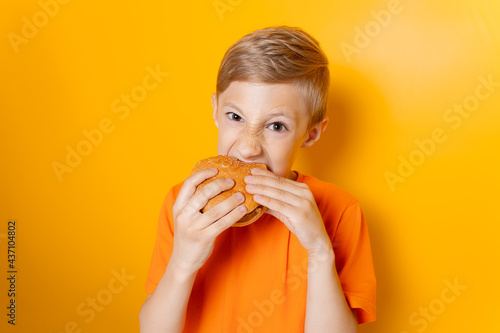 a boy in an orange T-shirt holds a hamburger in both hands and greedily bites it on a yellow background