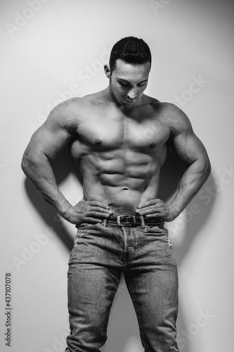 A young athlete bodybuilder poses in the studio topless in jeans near the wall. Black and white.