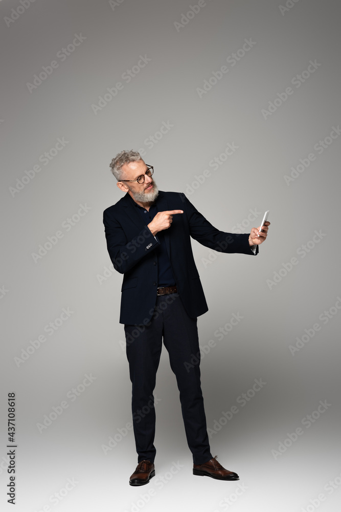 full length of businessman with grey hair pointing with finger while taking selfie on grey full length of businessman with grey hair pointing with finger while taking selfie on grey