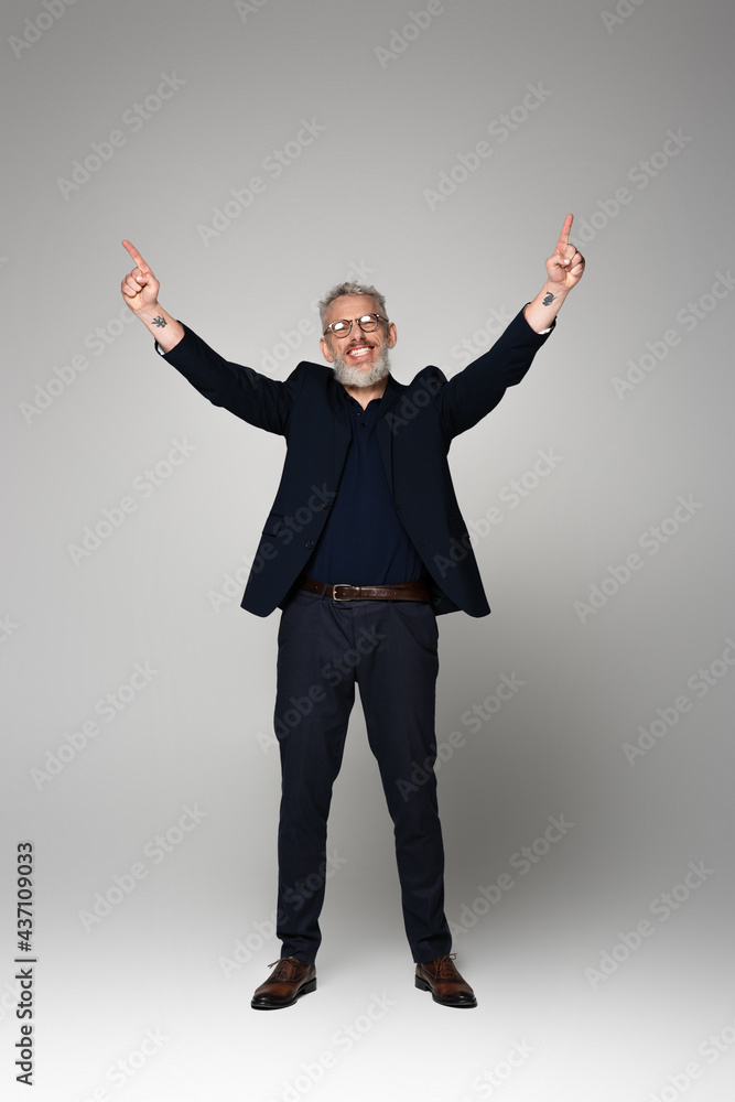 full length of smiling middle aged man in glasses and suit pointing with fingers on grey full length of smiling middle aged man in glasses and suit pointing with fingers on grey