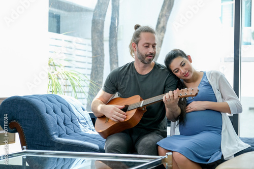 Pregnant woman sit beside and cuddle her husband who play guitar for his wife and they look happy to stay in living room at home together.