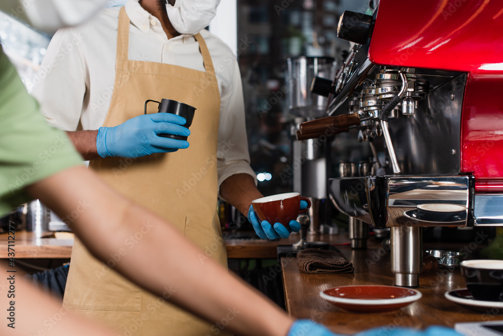 Cropped view of african american barista in medical mask and latex gloves holding cup and making coffee near blurred colleague