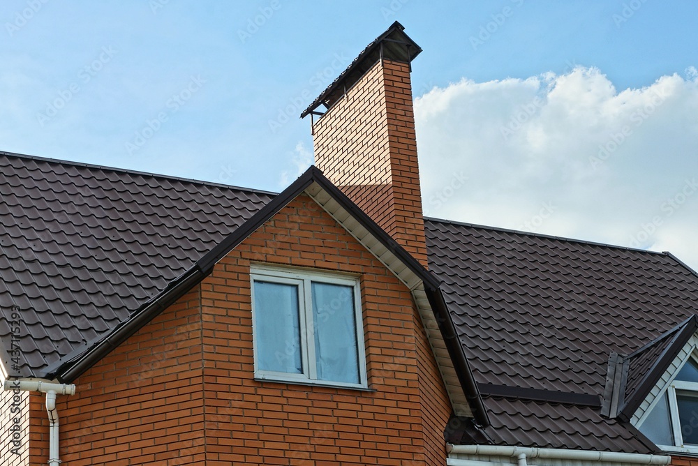 red brick attic with a white window and one large chimney on a brown tiled roof against a background of blue sky and clouds