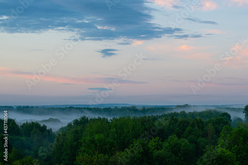 Fog spreads over the lowlands along the forest during sunset. Evening landscape. Clouds in the setting sky.