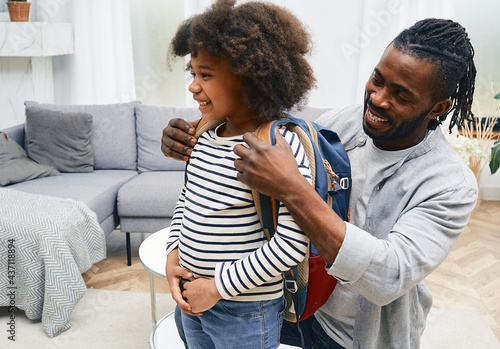 Happy African American daddy helps daughter getting ready for school and puts a school bag on her back photo