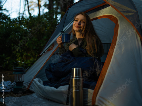 In the photo it is night. A young beautiful woman is sitting in the tent. She is holding a blue cup in her hands. In the foreground is a large thermos. In the left corner there is a photo-lamp.