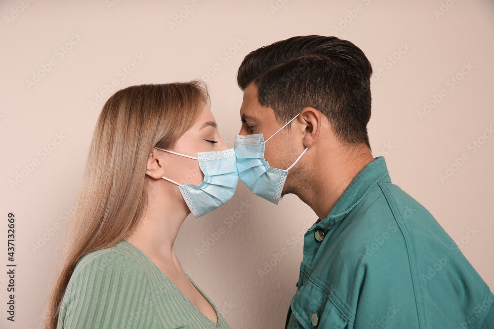 Couple in medical masks trying to kiss on beige background