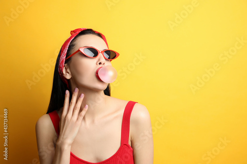 Fashionable young woman in pin up outfit blowing bubblegum on yellow background, space for text