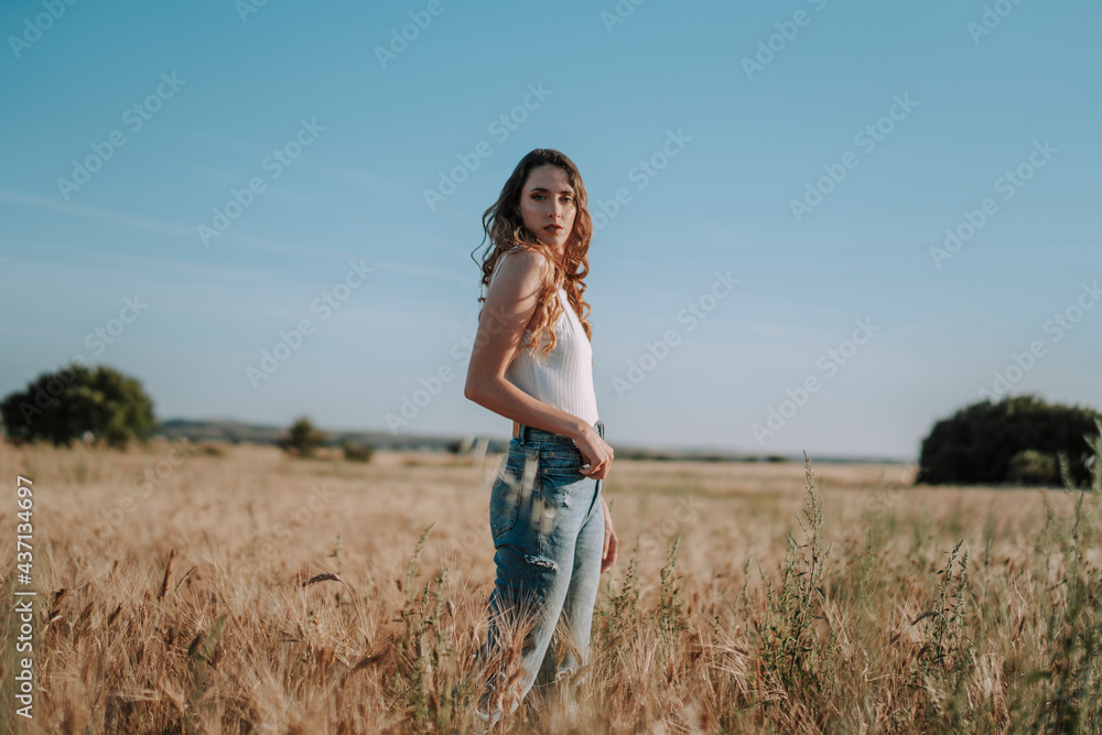 Chica joven atractiva posando en mitad de un campo de trigo al atardecer con tonos dorados Chica joven atractiva posando en mitad de un campo de trigo al atardecer con tonos dorados