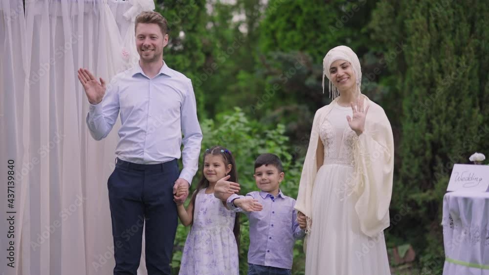 Happy newlyweds and children holding hands waving to guests on marriage ceremony. Cheerful Middle Eastern bride and Caucasian groom with boy and girl standing at wedding altar outdoors smiling Happy newlyweds and children holding hands waving to guests on marriage ceremony. Cheerful Middle Eastern bride and Caucasian groom with boy and girl standing at wedding altar outdoors smiling