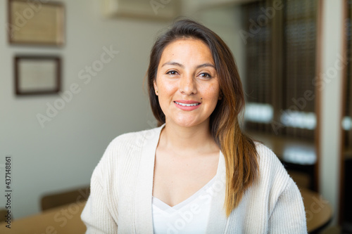 Closeup portrait of young smiling brown-haired hispanic woman wearing light knitted jacket standing on blurred background of office meeting room.