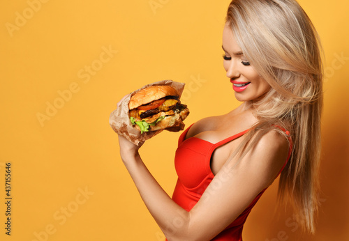 Pretty sexy smiling blonde woman in red swimsuit with deep neckline holding and looking at fresh cheeseburger in hand over yellow wall background. Diet, slimming, fast food, weight loss concept