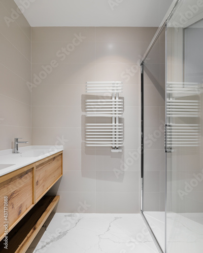 Modern bathroom with wooden base for white sinks. Radiator for warm water procedures in winter. Shower zone with glass door. Bathroom decorated with light tiles