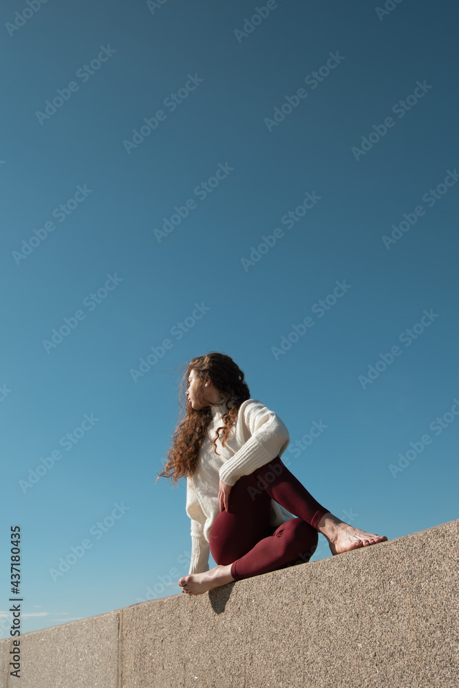 Young brunette woman practicing Ardha Matsyendrasana pose on top of concrete wall on a beach in front of blue sky Young brunette woman practicing Ardha Matsyendrasana pose on top of concrete wall on a beach in front of blue sky