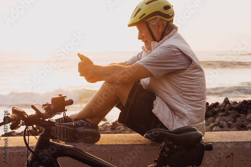 Cyclist man using mobile phone sitting along the sea at sunset, close his bike. Retired people enjoying healthy lifestyle